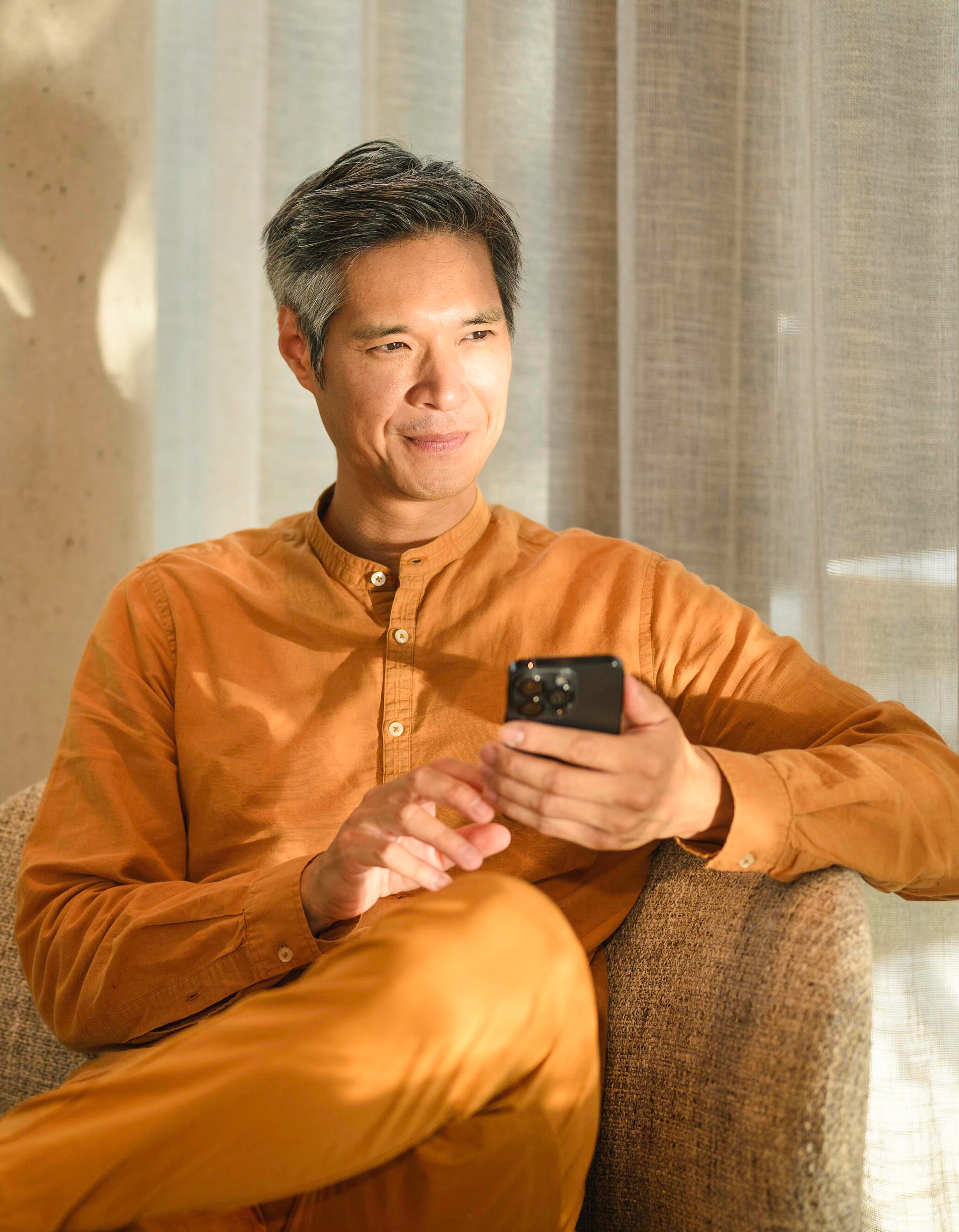 A person sitting on a sofa, holding a mobile device and looking into the distance.