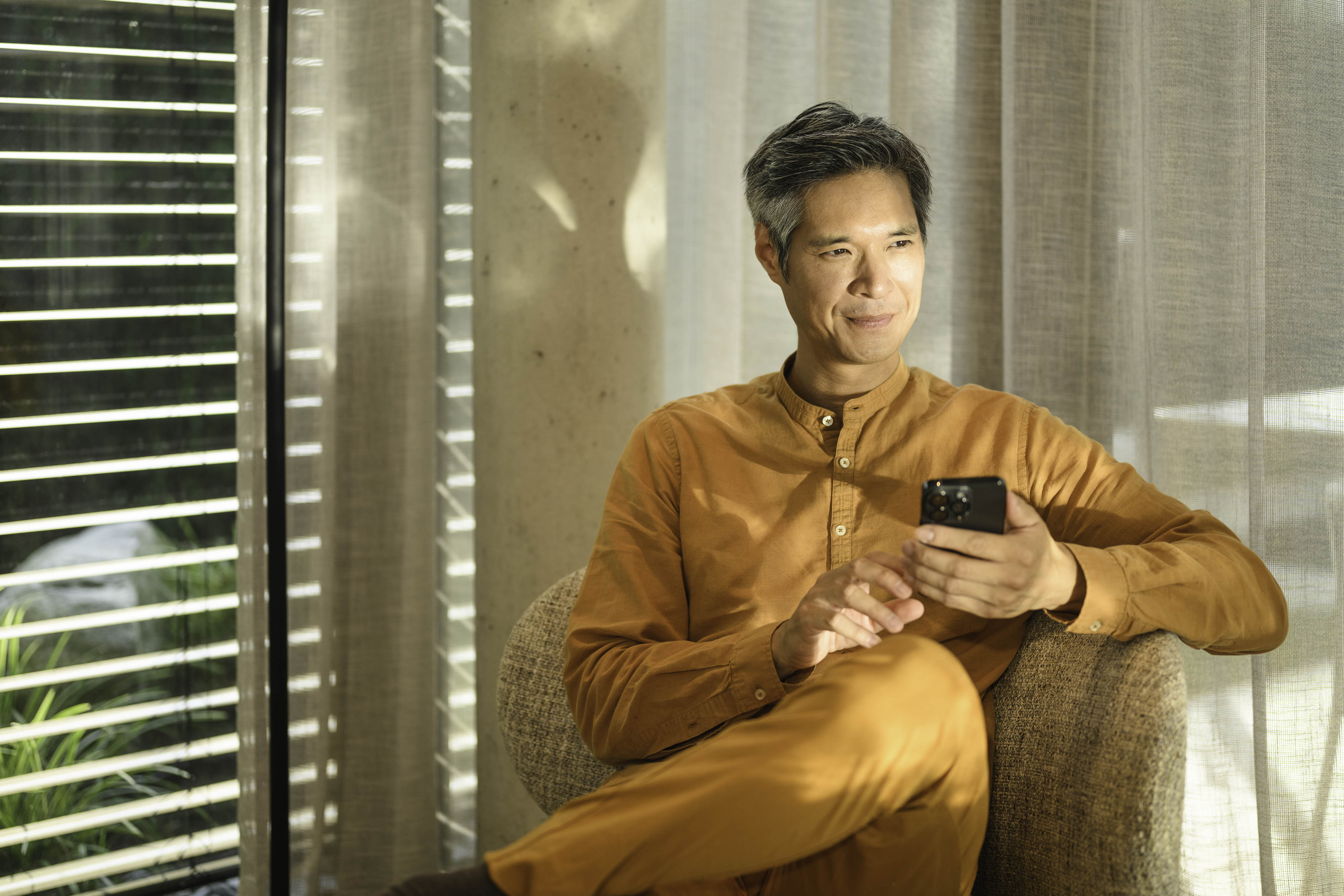 A person sitting on a sofa, holding a mobile device and looking into the distance. Online access to existing customers account