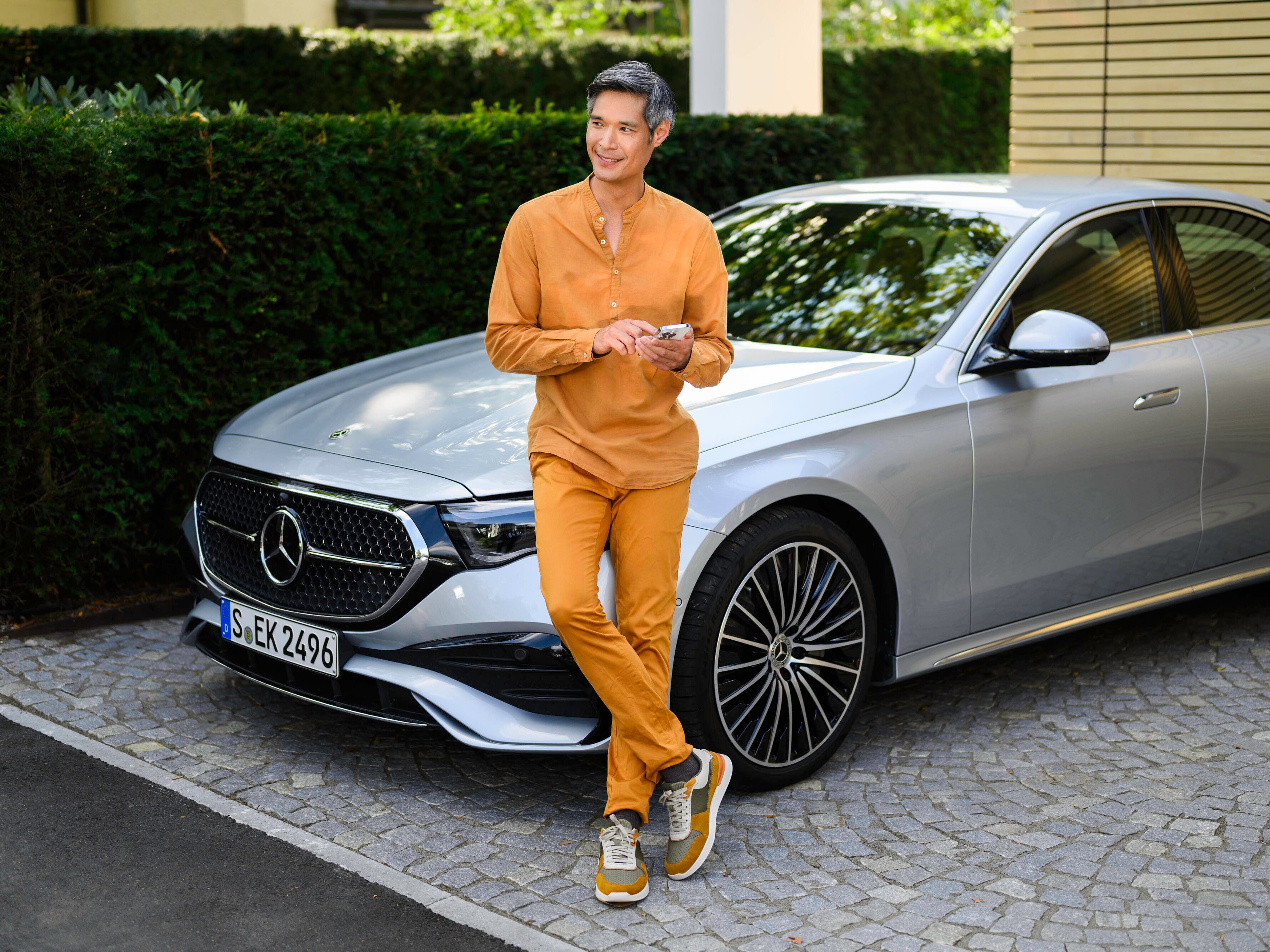 A person leaning against a Mercedes-Benz vehicle, looking at distance and using a mobile device. 