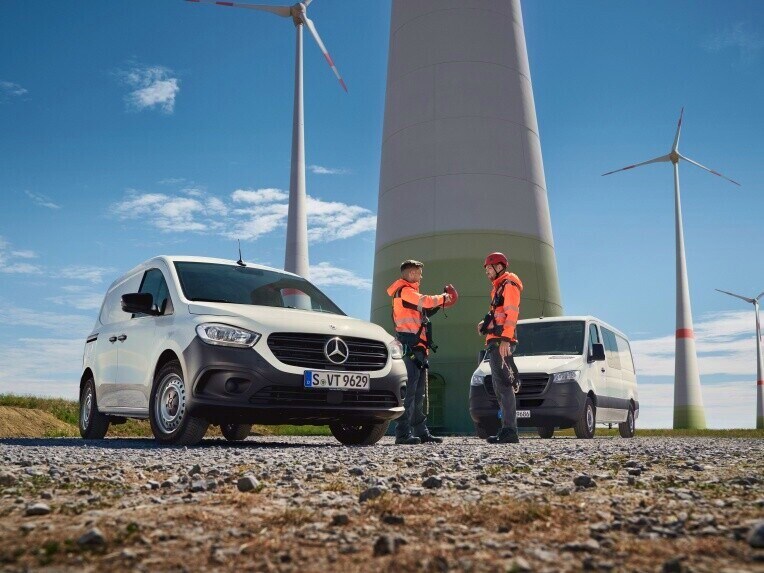 Two Mercedes-Benz vans and two people in high-visibility clothing in front of a wind farm.