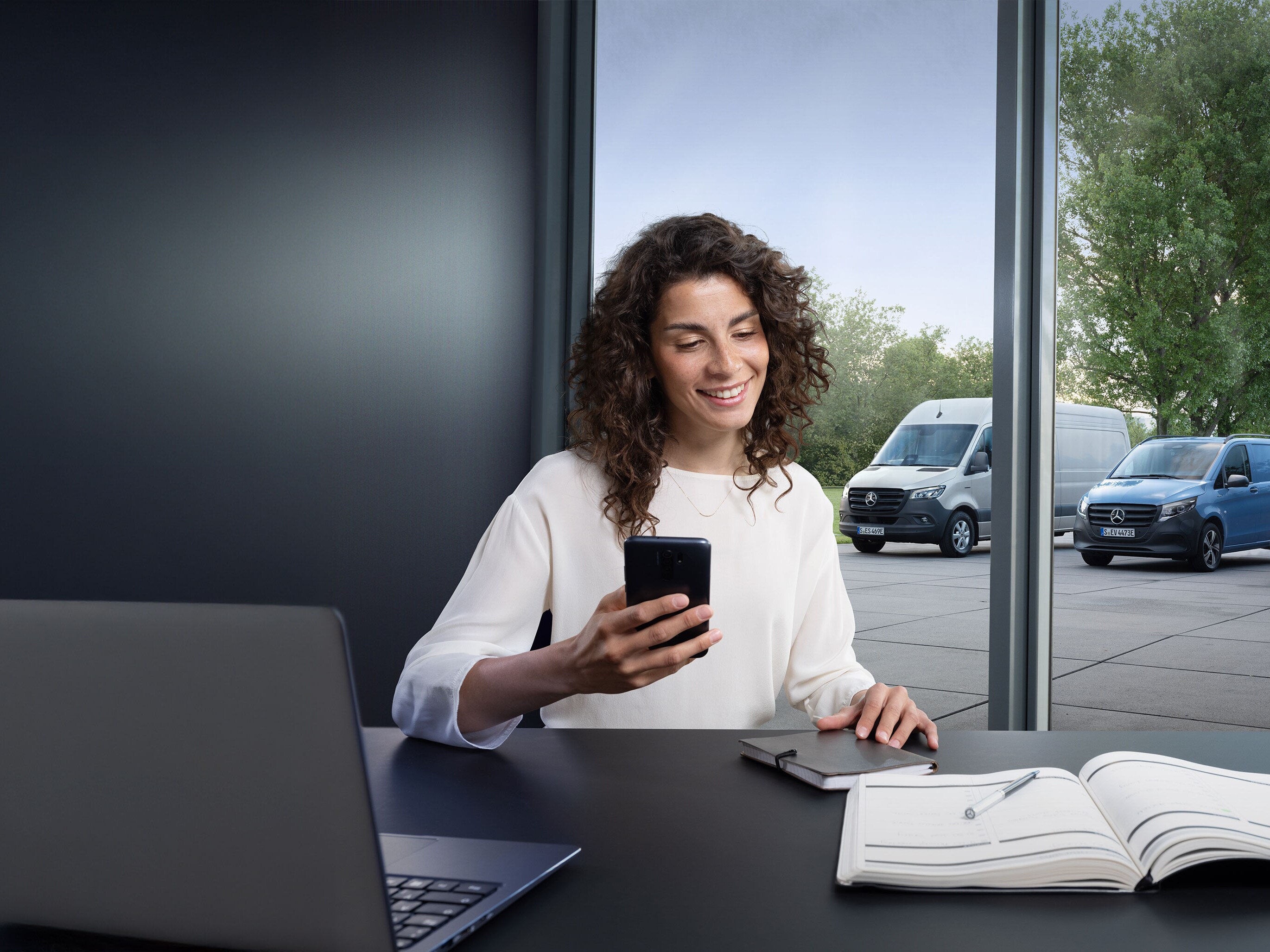 Woman booking a service via smartphone with a range of Mercedes-Benz vans in the background.