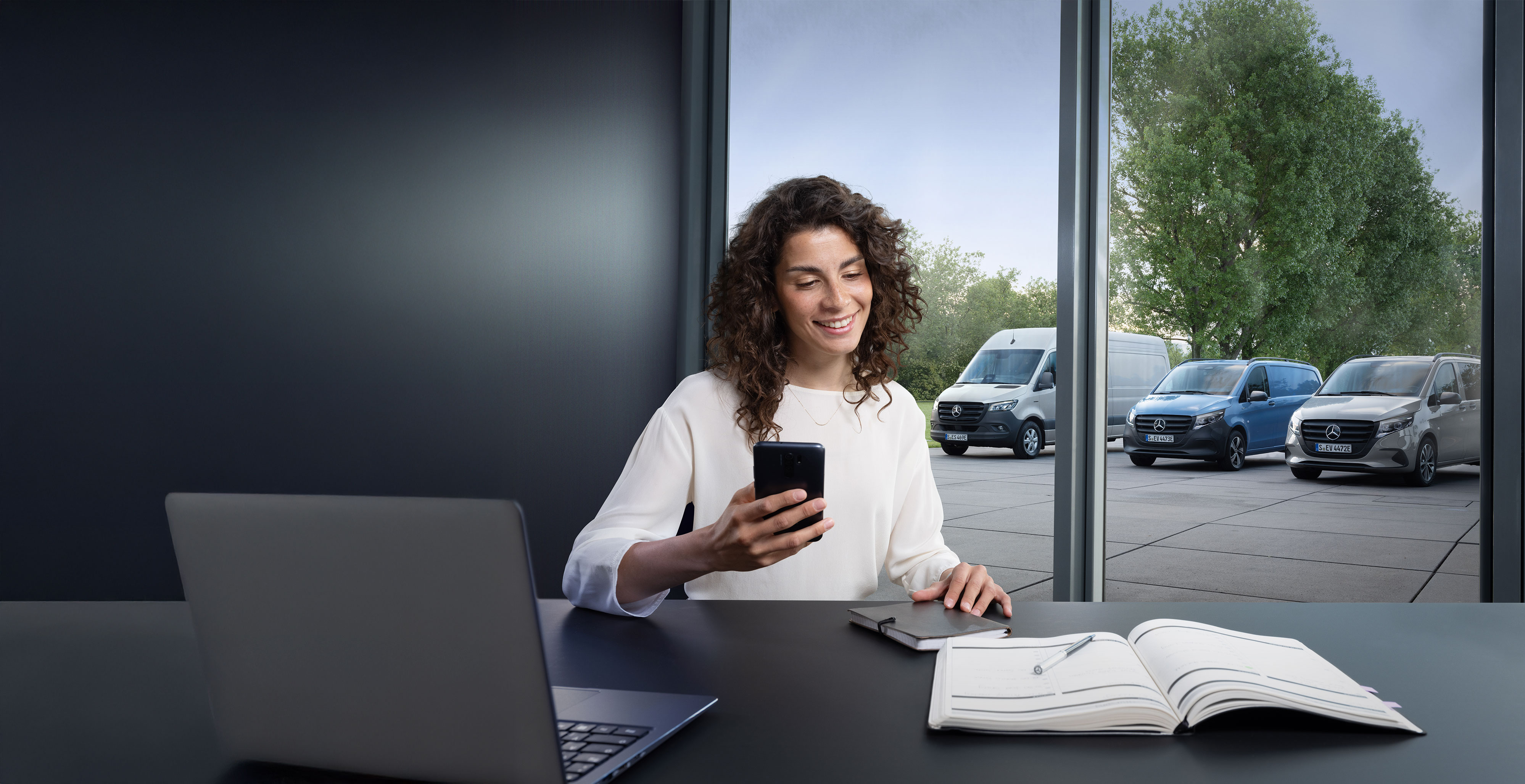 Mercedes-Benz | Convenience | Online Appointment Booking Woman booking a service via smartphone with a range of Mercedes-Benz vans in the background.