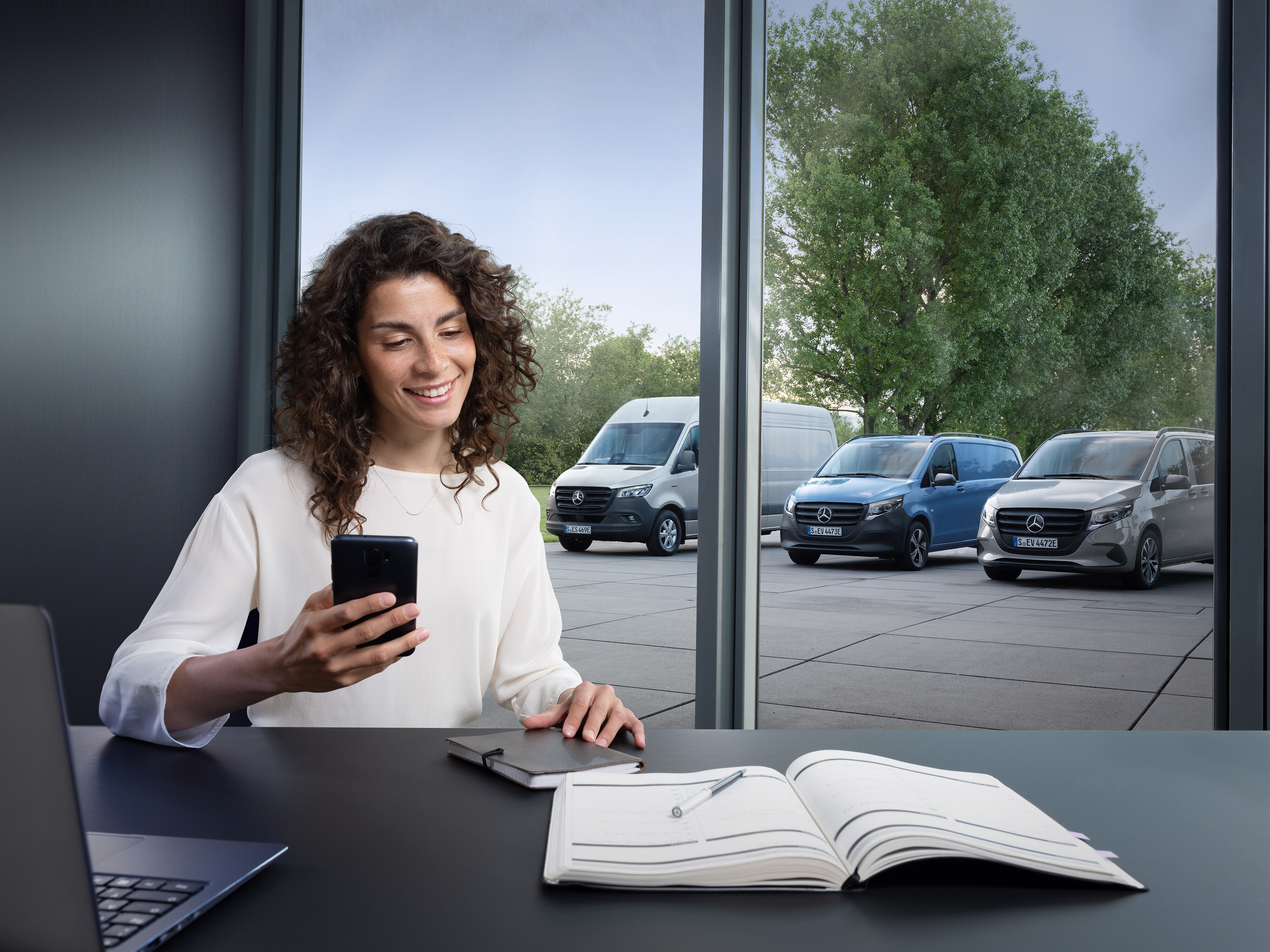 Woman with a smartphone in front of a window with three Mercedes-Benz vans standing outside.