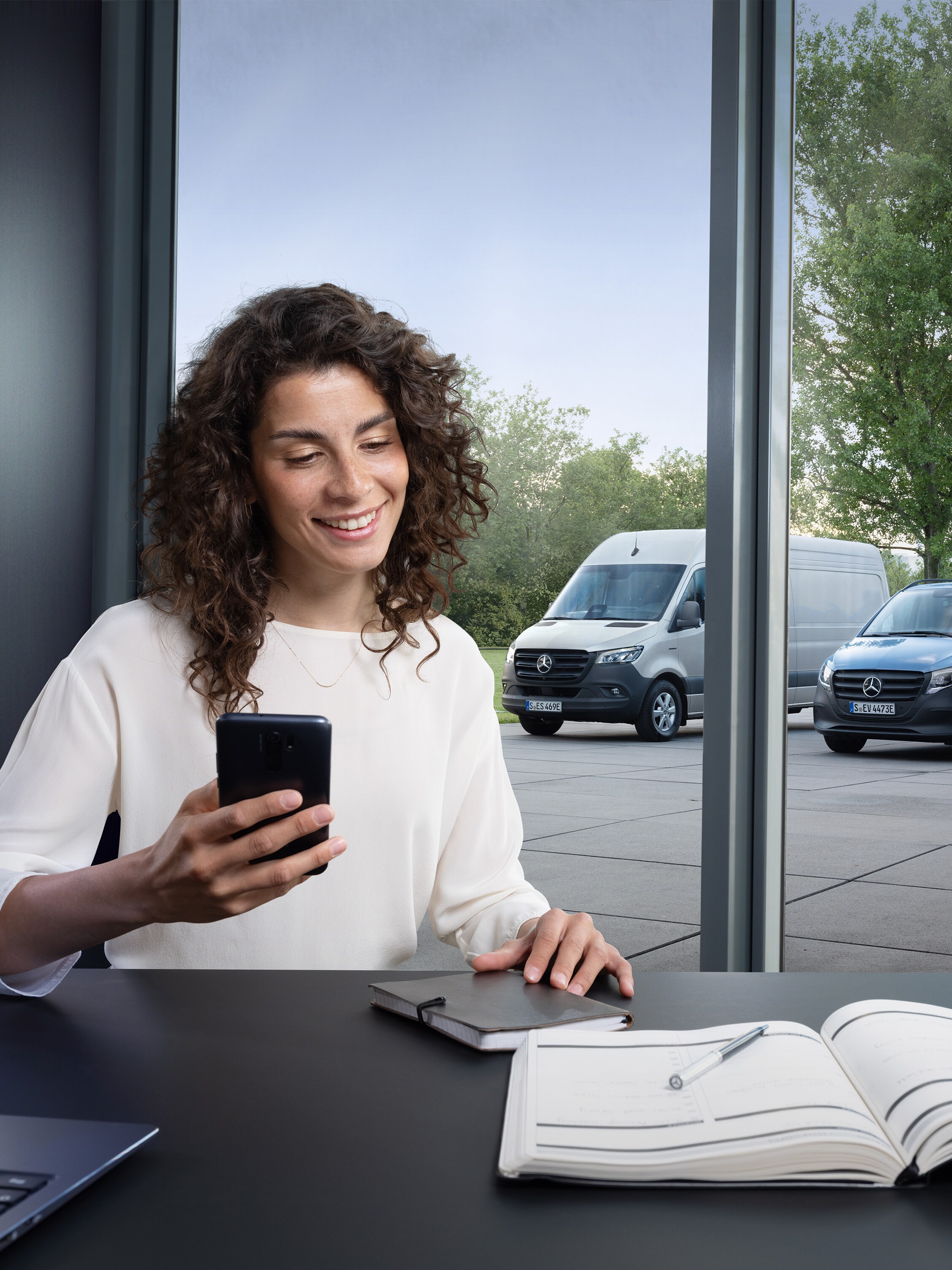 Woman with a smartphone in front of a window with three Mercedes-Benz vans standing outside.