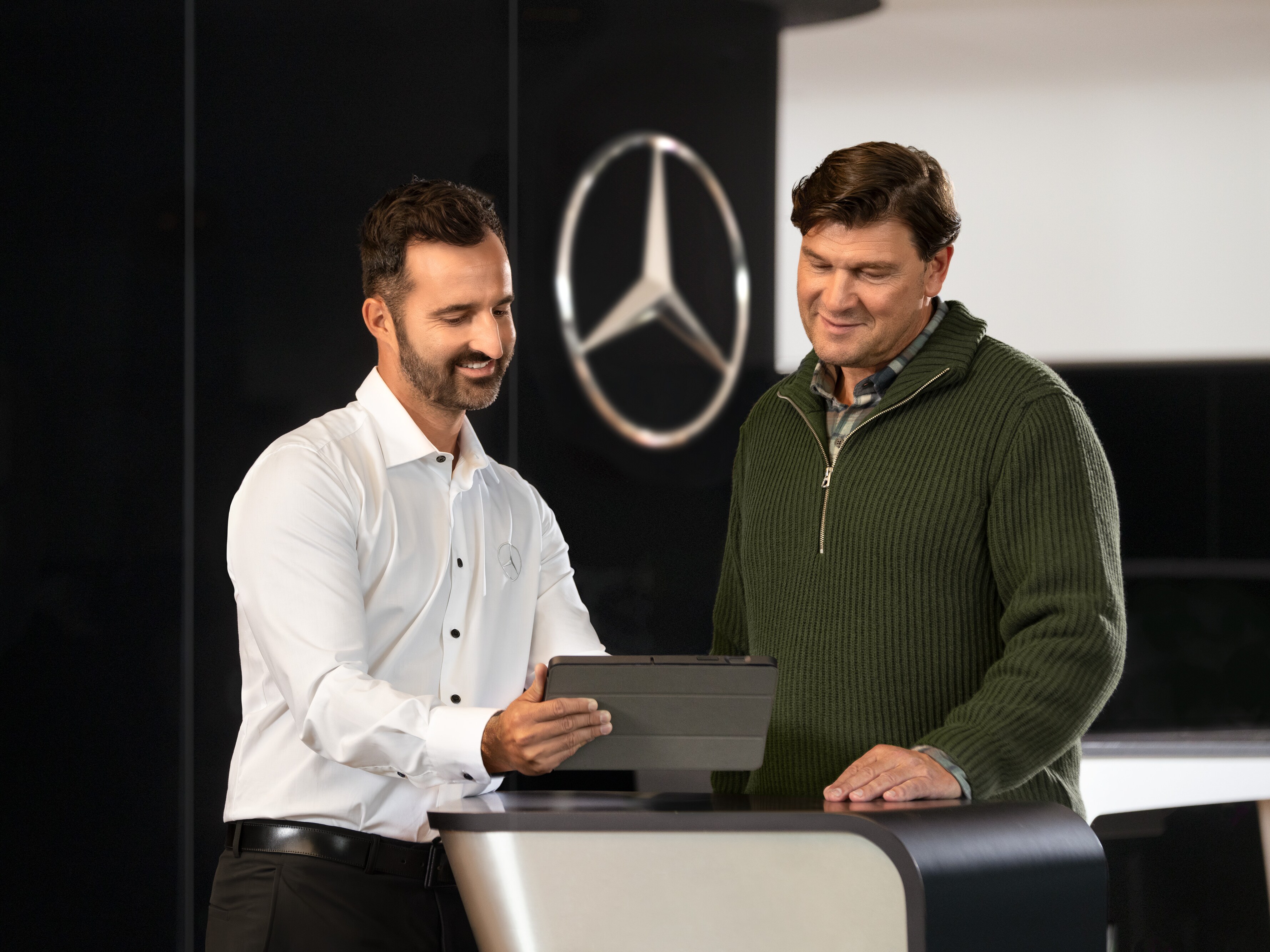Two men talking at a counter, while looking at a tablet, in front of the Mercedes-Benz star.